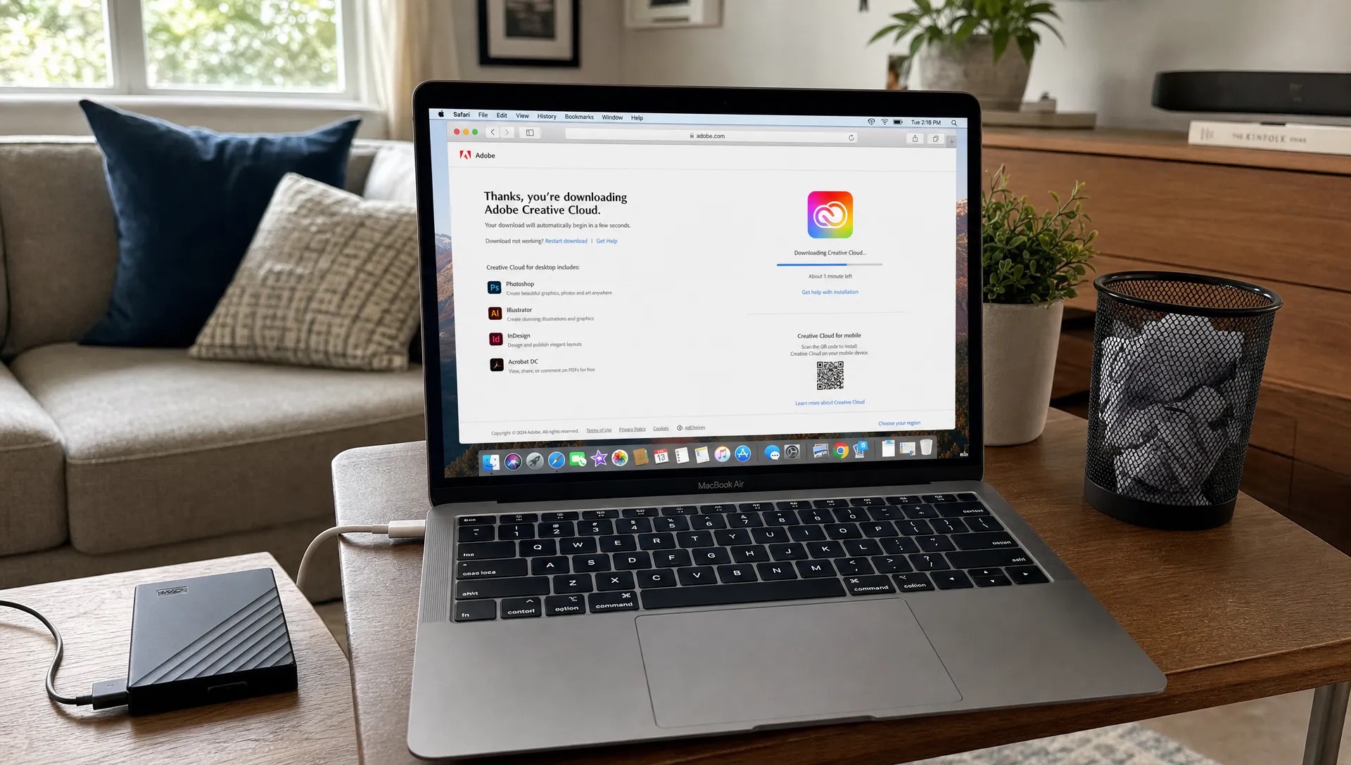 A Mac laptop in a living room work corner shows a download page beside an external drive and trash can.